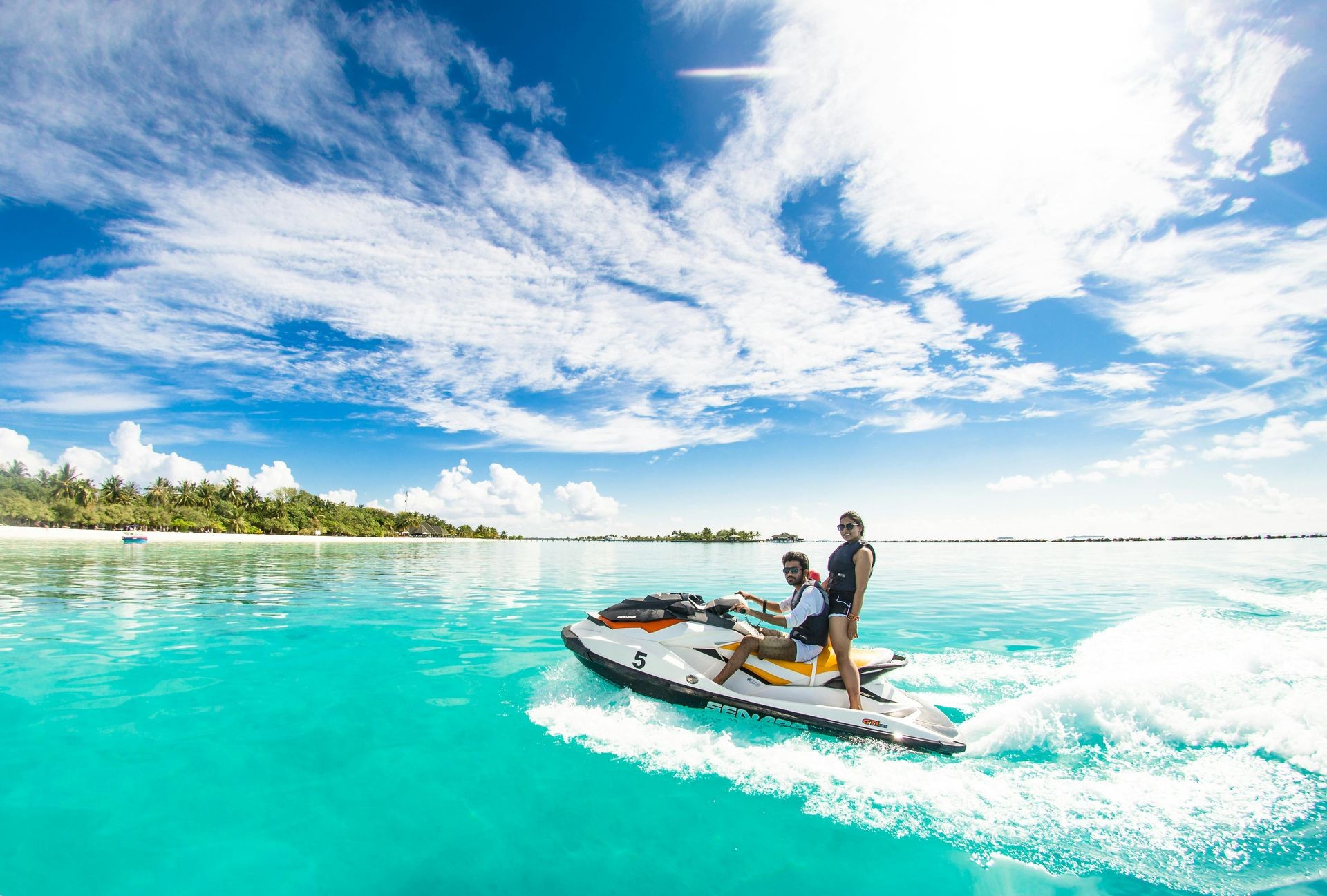 Two people riding a jet ski on clear turquoise water with a sunny blue sky and distant shoreline.